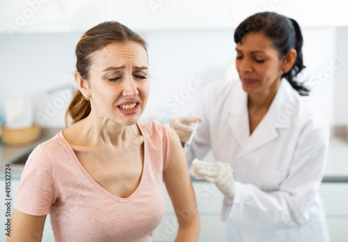 Hispanic woman doctor vaccinating young caucasian woman patient who writhing in pain.