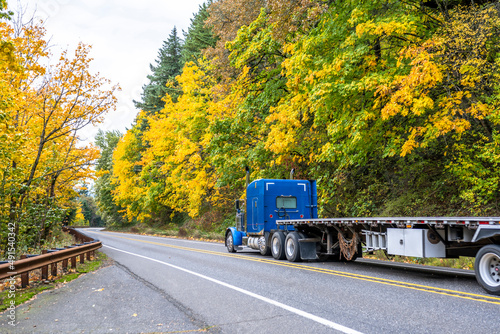 Commercial classic blue big rig semi truck with empty flat bed semi trailer running on the winding mountain road with yellow autumn trees on the slopes