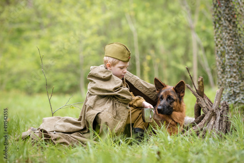 a boy in a military uniform in a clearing, sitting by a campfire with a German shepherd.Two friends defend the motherland
