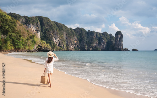 Rear view image of a young woman with hat and bag walking on the beach with blue sky background