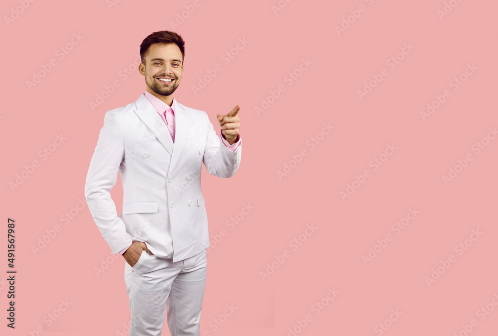 Smiling man in white formal suit looks at you and points his index finger at copy space on pink background. Happy young attractive caucasian man advertises or recommends. Creative banner. Smiling man in white formal suit looks at you and points his index finger at copy space on pink background. Happy young attractive caucasian man advertises or recommends. Creative banner.