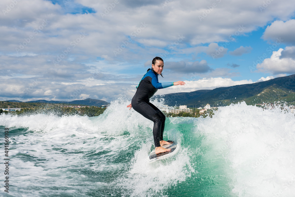 Young athletic female in wetsuit riding on endless waves behind a boat on sunny day. Woman practice wakeboarding, carving behind the baot. Watersport concept. Young athletic female in wetsuit riding on endless waves behind a boat on sunny day. Woman practice wakeboarding, carving behind the baot. Watersport concept.