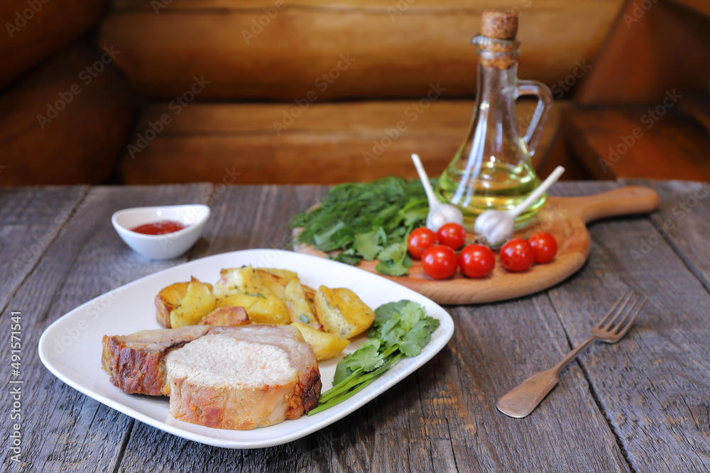 On a white ceramic plate are a big chunk of baked pork with rustic fried potatoes on the background of a wooden cutting board with tomatoes, herbs and a jug of oil. 