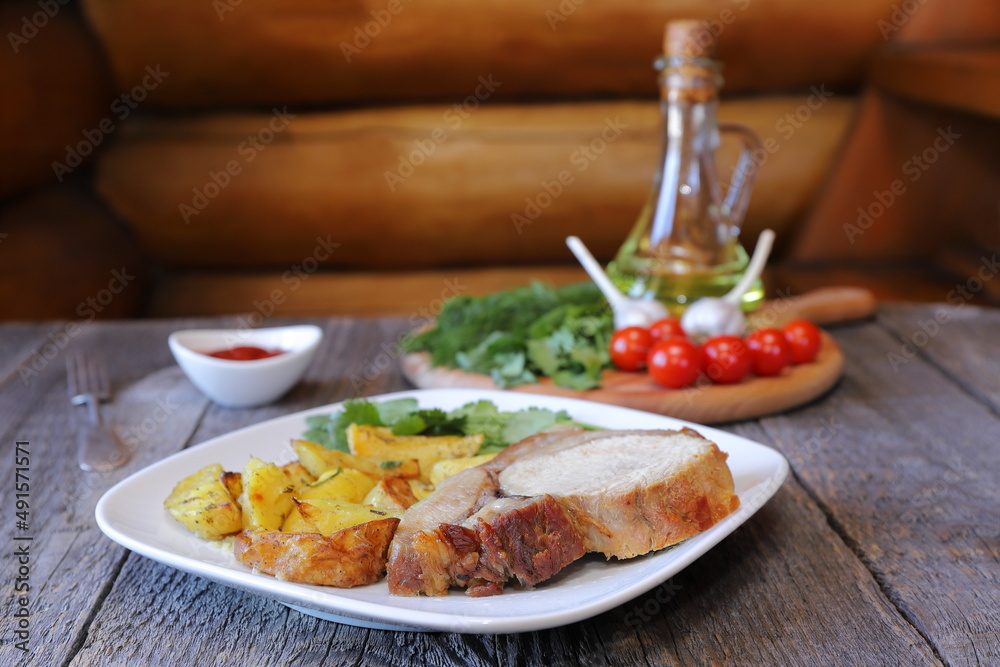 On a white ceramic plate are a big chunk of baked pork with rustic fried potatoes on the background of a wooden cutting board with tomatoes, herbs and a jug of oil. 