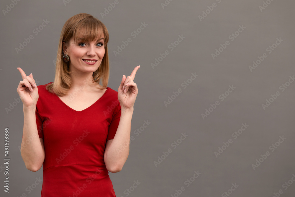 Happy surprised blonde young female smiling broadly at camera, pointing fingers away, showing something interesting and exciting on studio wall with copy space for your text or advertising content Happy surprised blonde young female smiling broadly at camera, pointing fingers away, showing something interesting and exciting on studio wall with copy space for your text or advertising content