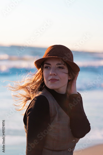 Beautiful girl posing with hat at beach. She is looking back. Sea background. Her red hair is flying with wind. 