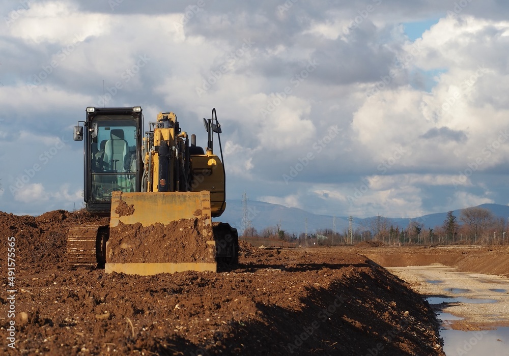 Front view of an excavator, with the lighted empty cabin, between a cloudy sky and the dirt soil of a new urban development site Front view of an excavator, with the lighted empty cabin, between a cloudy sky and the dirt soil of a new urban development site