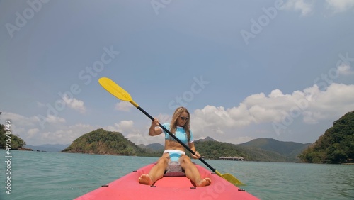 Pretty sexy young blonde woman in stylish swimsuit and sunglasses enjoys trip canoe on sea against palm islands. Traveling to tropical countries. Positive sport girl hand padding on kayak front view.