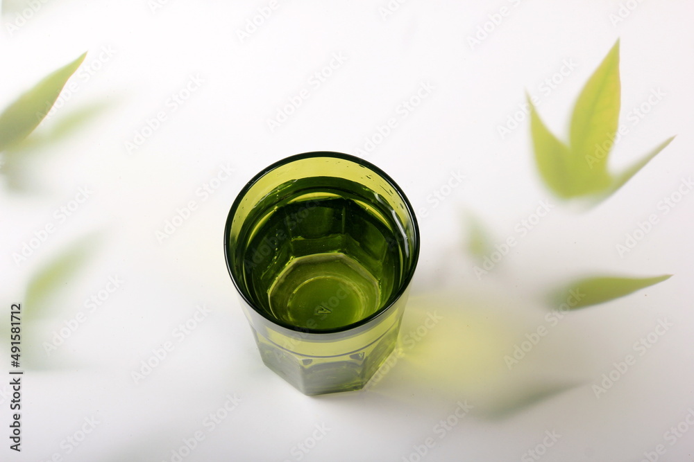 Green glass with water and a light background with plant leaves. Top view green glass of water with sunlight long shadow and refraction patterns and palm leaf trendy shadow 