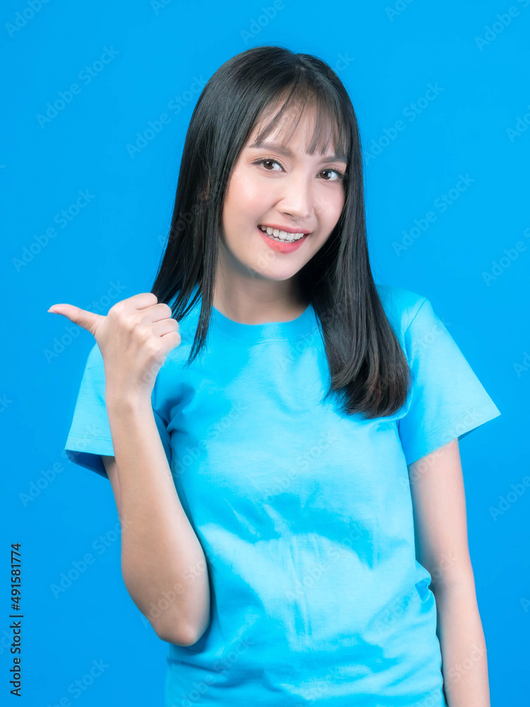 Beautiful Asian woman cute girl with bangs hair style in blue t shirt smiling and pointing finger to empty copy space for present product or blank space for advertising isolated on blue background Beautiful Asian woman cute girl with bangs hair style in blue t shirt smiling and pointing finger to empty copy space for present product or blank space for advertising isolated on blue background
