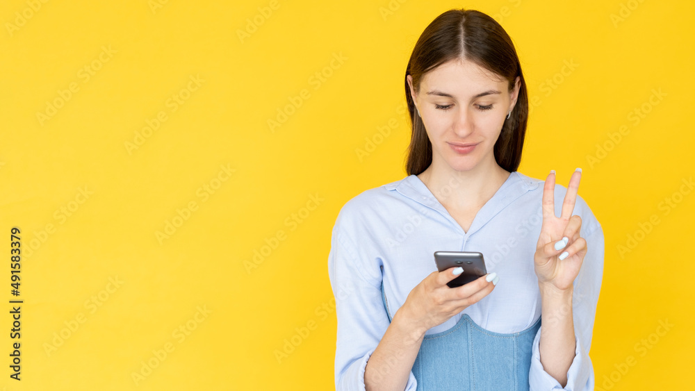 Online communication. Positive woman. Peace gesture. Smiling pretty lady showing victory sign typing smartphone isolated yellow copy space. Online communication. Positive woman. Peace gesture. Smiling pretty lady showing victory sign typing smartphone isolated yellow copy space.