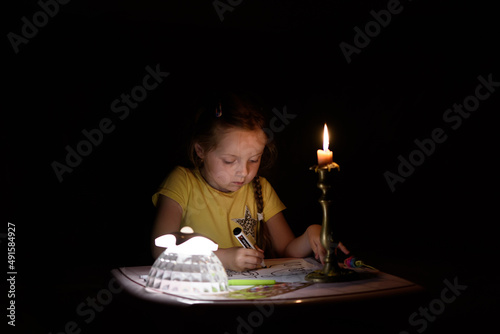 Little girl draws fantasy characters near candle in a dark room. Child doing favorite thing during a power outage