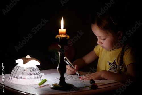 Little girl draws fantasy characters near candle in a dark room. Child doing favorite thing during a power outage