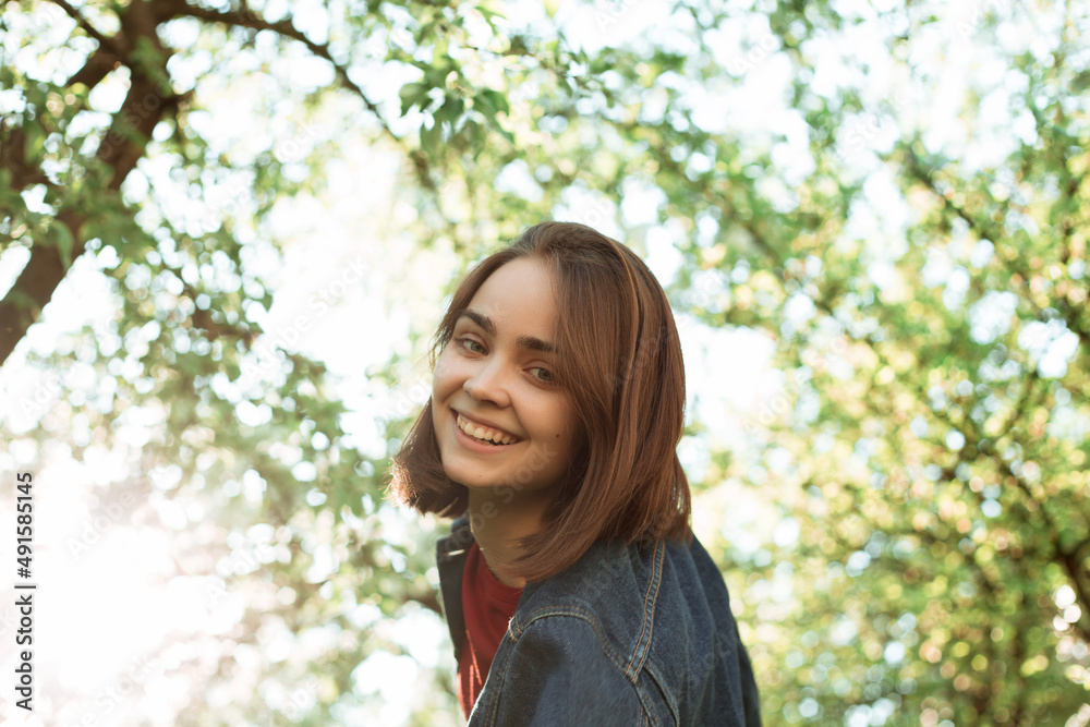 From below of positive young woman in denim jacket standing in green park against trees and looking at camera From below of positive young woman in denim jacket standing in green park against trees and looking at camera