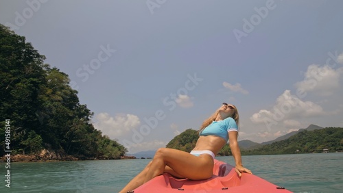 фотографія Pretty young blonde woman in stylish swimsuit and sunglasses sits on pink canoe sailing along sea against hills