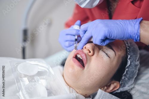 Cosmetologist doing painful lip augmentation procedure with hyaluronic acid. The beautician pierces lips by needle. Woman suffering subcutaneous injection to increase lips shape with dermal filler.