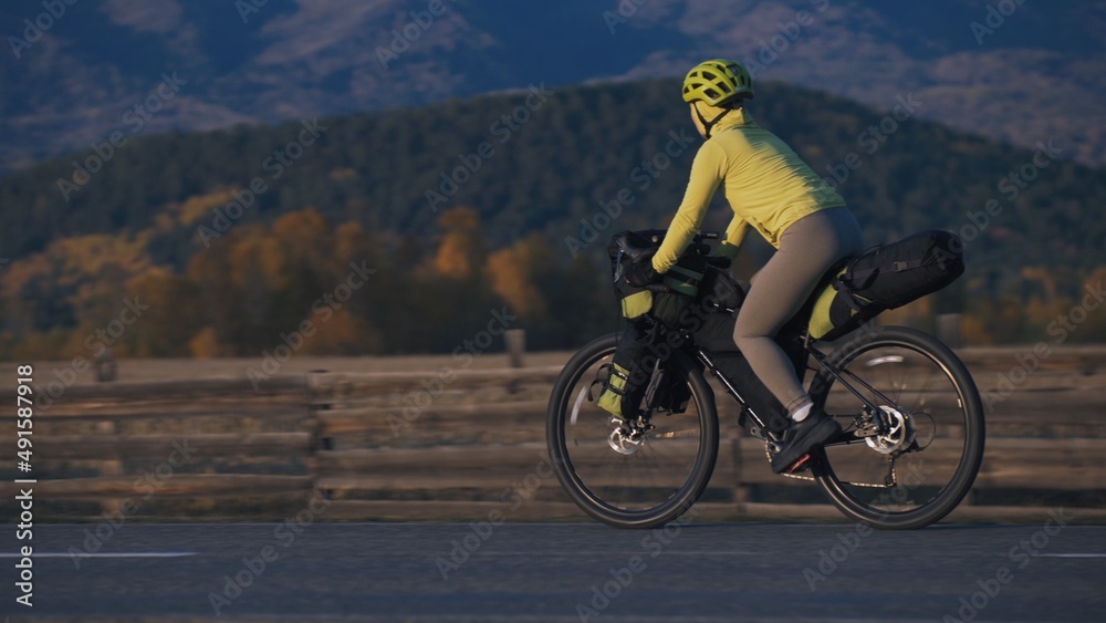 Fototapeta premium The woman travel on mixed terrain cycle touring with bikepacking. The traveler journey with bicycle bags. Sport tourism bikepacking, bike, sportswear in green black colors. Mountain snow capped.