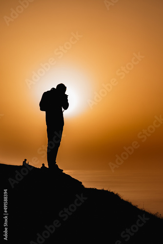 Silhouette of a man taking pictures from a cliff watching the ocean with the sunset in the background. © MartiFerretPhoto