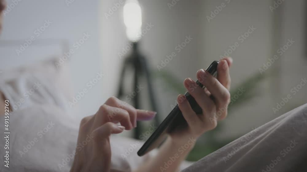Unrecognizable woman uses smartphone while lying in bedroom. Hands and phone close up.