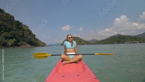 Attractive sportive blonde woman with sunglasses rows pink plastic canoe along sea water against green hills and blue sky. Traveling to tropical countries. Sports girl is sailing on kayak in ocean. photo