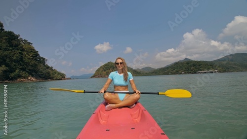 Attractive sportive blonde woman with sunglasses rows pink plastic canoe along sea water against green hills and blue sky. Traveling to tropical countries. Sports girl is sailing on kayak in ocean. © ivandanru