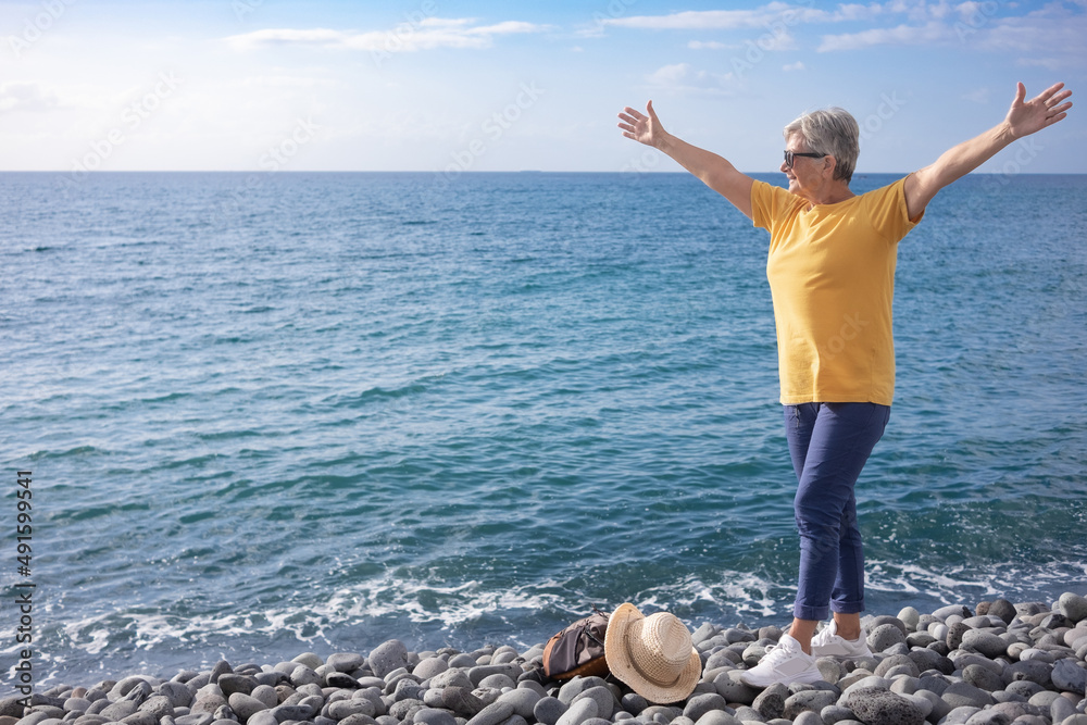 Senior caucasian woman standing with open arms on a pebble beach at sea looking at horizon over water. Concept of vacation, freedom and happy retirement