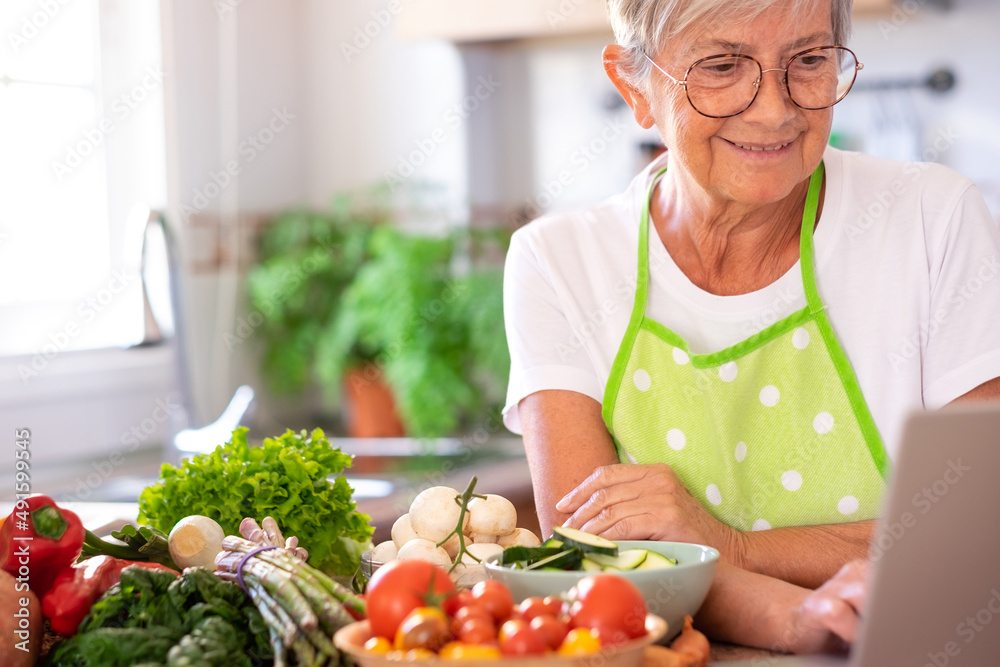 Attractive senior woman preparing vegetables in the home kitchen searching menu at laptop computer. Caucasian elderly people enjoying healthy eating Attractive senior woman preparing vegetables in the home kitchen searching menu at laptop computer. Caucasian elderly people enjoying healthy eating
