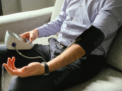 Man is checking blood pressure at home. Sitting on a grey sofa. Monitoring of Seasonal health issues or stress at work Hypertension or hypertension seasonal disorder and problems Taking care of health