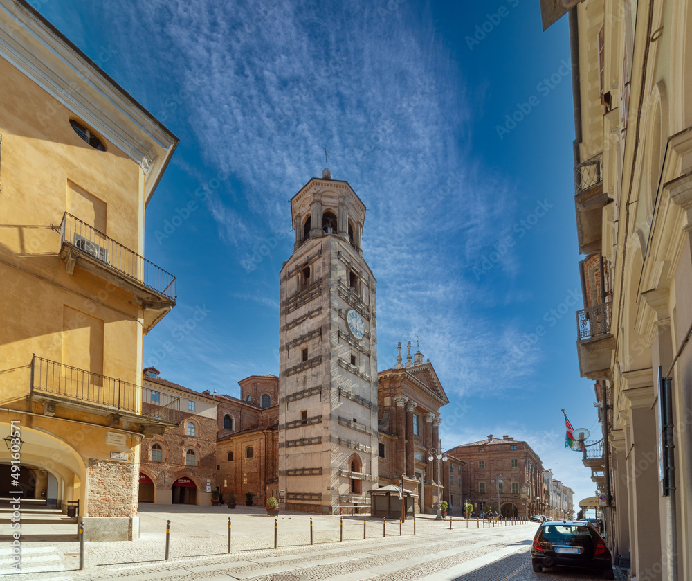 Fototapeta premium Fossano, Italy - February 27, 2022: the Civic bell tower (XV century) and Cathedral of Santa Maria and San Giovenale (XVIII century) with historic buildings in via Roma
