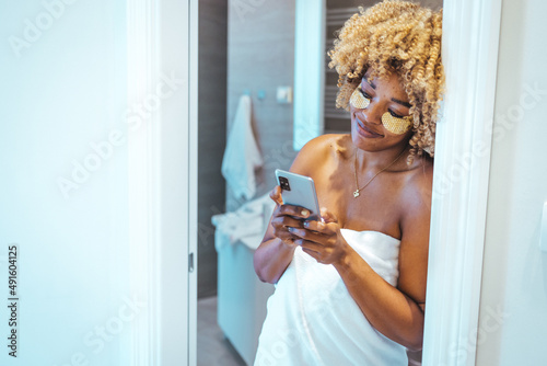 Woman in the bathroom with mobile phone. Mixed race woman wearing bathrobe using smartphone in bathroom. Happy Woman Talking On Phone Applying Patches In Bathroom