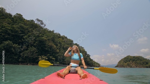 Pretty young blonde woman in stylish swimsuit and sunglasses sits on pink canoe sailing along sea against hills. Traveling to tropical countries. Positive sport girl hand padding on kayak front view.
