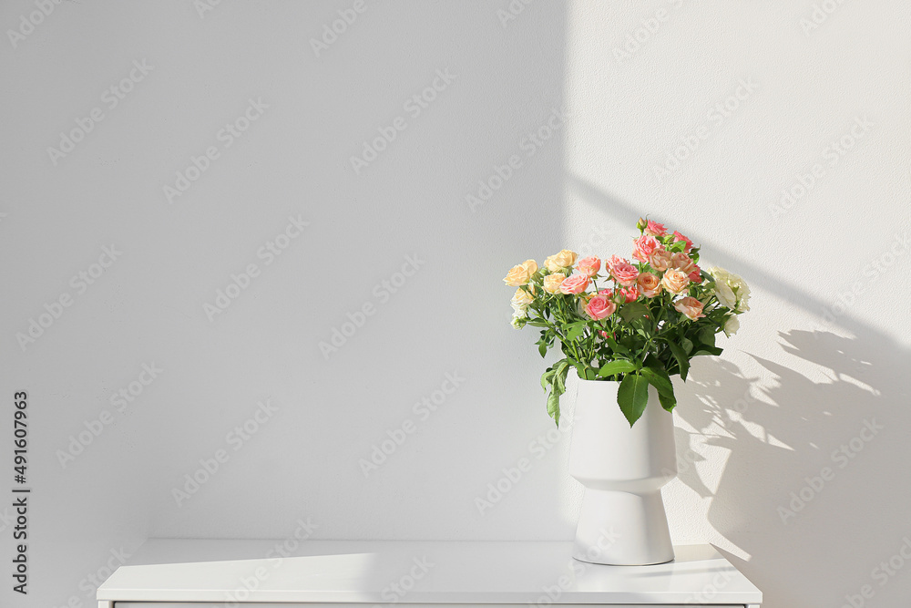 White vase with beautiful roses on shelf near light wall