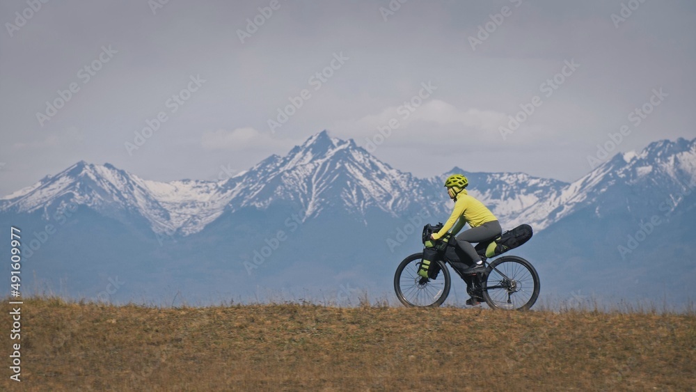 The woman travel on mixed terrain cycle touring with bikepacking. The traveler journey with bicycle bags. Sport tourism bikepacking, bike, sportswear in green black colors. Mountain snow capped.