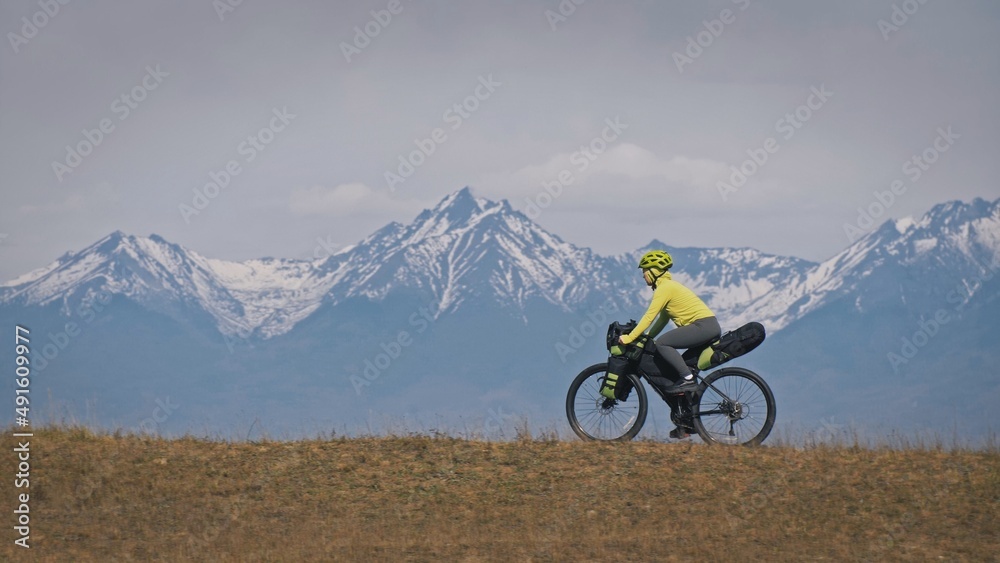 Obraz premium The woman travel on mixed terrain cycle touring with bikepacking. The traveler journey with bicycle bags. Sport tourism bikepacking, bike, sportswear in green black colors. Mountain snow capped.