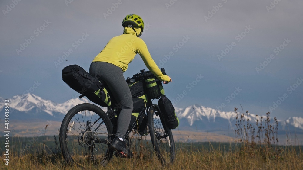 The woman travel on mixed terrain cycle touring with bikepacking. The traveler journey with bicycle bags. Sport tourism bikepacking, bike, sportswear in green black colors. Mountain snow capped. The woman travel on mixed terrain cycle touring with bikepacking. The traveler journey with bicycle bags. Sport tourism bikepacking, bike, sportswear in green black colors. Mountain snow capped.