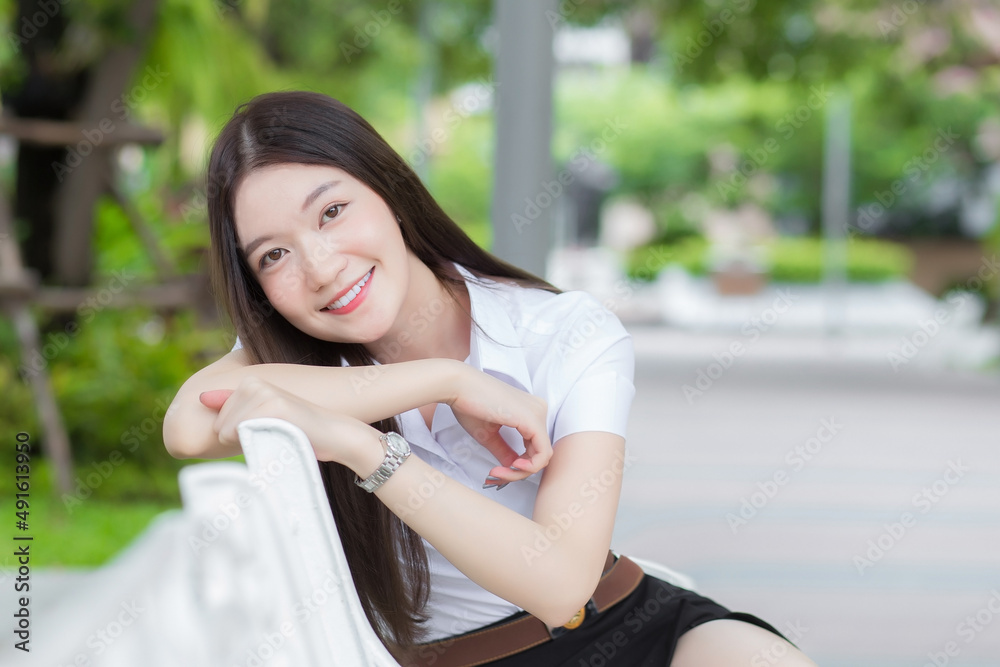 Portrait of an adult Thai student in university student uniform. Asian beautiful girl sitting smiling happily at university with a background of garden trees. Portrait of an adult Thai student in university student uniform. Asian beautiful girl sitting smiling happily at university with a background of garden trees.