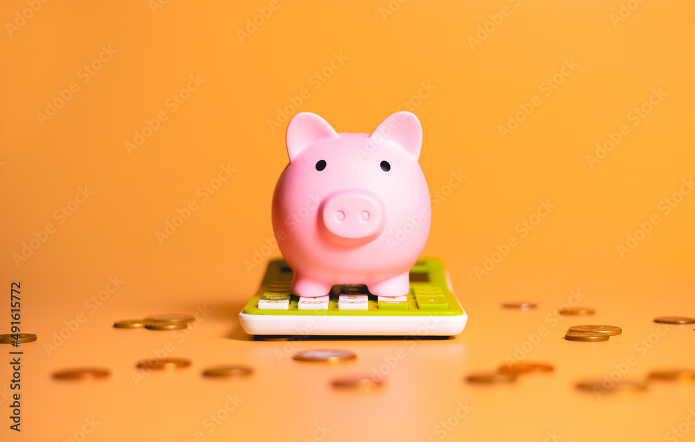 A piggy bank over a green calculator with scattered coins isolated on orange background in studio photography. Concepts of saving money, finance and investments. - obrazy, fototapety, plakaty 