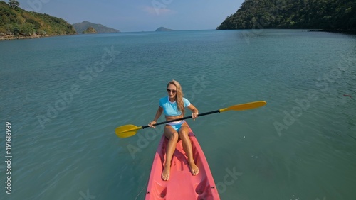Pretty sportive blonde lady in stylish swimsuit sails on pink plastic canoe along azure ocean bay at exotic resort. Traveling to tropical countries. Girl is sailing on kayak in ocean, view from above.