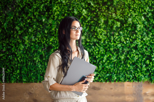 Portrait modern female woman using laptop and phone, communicates on internet with customer in city cafe. Cozy office workplace. Eco-friendly background wall of green grass. Copy space
