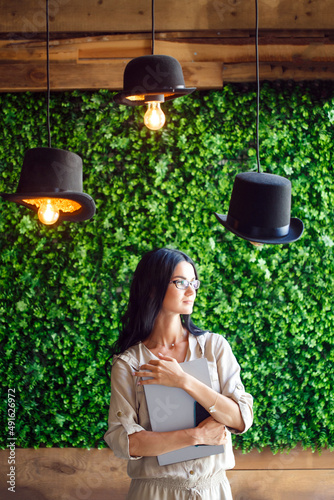 Portrait modern female woman using laptop and phone, communicates on internet with customer in city cafe. Cozy office workplace. Eco-friendly background wall of green grass