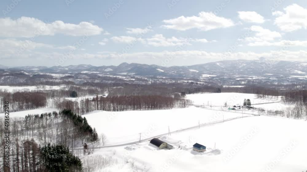 Large farms and a large barn near the arable fields completely covered with a thick layer of snow on a partly cloudy day in Kutchan in Japan. High angle drone dolley shot Large farms and a large barn near the arable fields completely covered with a thick layer of snow on a partly cloudy day in Kutchan in Japan. High angle drone dolley shot
