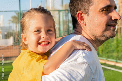 Side view of smiling father carrying cheerful girl on back while spending time together at football field