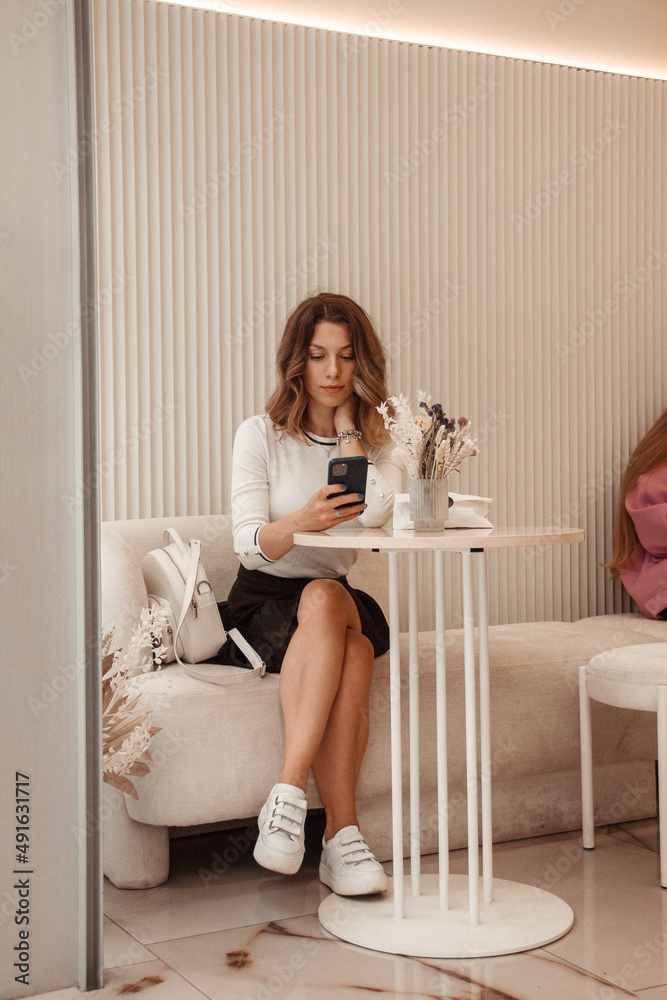 young curly brunette girl in white shirt and black skirt is sitting in beige modern cafe with mobile phone in hand and waiting for an order on the white wall background. lifestyle concept, free space
