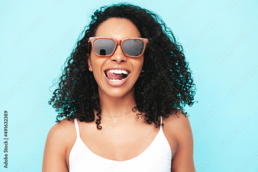 Beautiful black woman with afro curls hairstyle. Smiling model dressed in white summer dress. Sexy carefree female posing near blue wall in studio. Tanned and cheerful. In sunglasses. Shows tongue Beautiful black woman with afro curls hairstyle. Smiling model dressed in white summer dress. Sexy carefree female posing near blue wall in studio. Tanned and cheerful. In sunglasses. Shows tongue