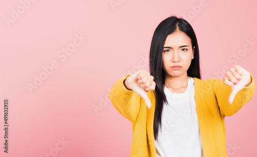 Portrait Asian beautiful young woman unhappy, negative gesture showing finger thumbs down or dislike sign, studio shot isolated on pink background, Thai female rejection unlike with copy space