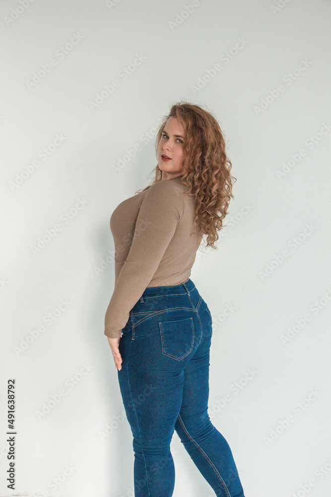 young plus size girl with red curly hair is standing from the back in the beige bodysuit and blue jeans and smiling cute on the white wall background. fashion concept, free space