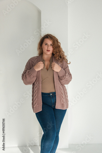 young plus size girl with red curly hair is standing in the beige cardigan and blue jeans and smiling cute on the white wall background. fashion concept, free space