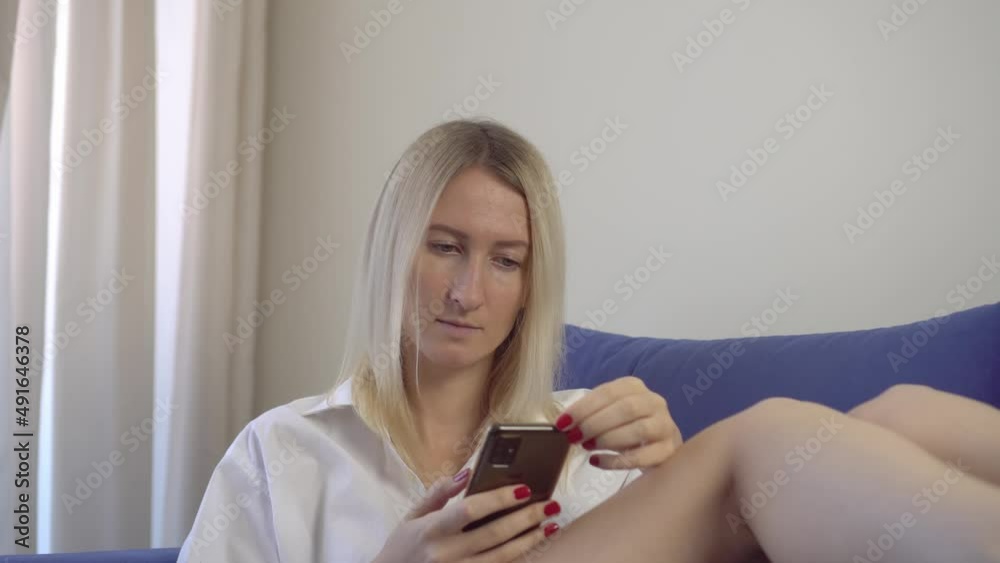 A relaxed woman at home reads a text message on her smartphone in her bright bedroom or hotel room. She's wearing a white shirt and jeans. Online communication. An application or messenger. A relaxed woman at home reads a text message on her smartphone in her bright bedroom or hotel room. She's wearing a white shirt and jeans. Online communication. An application or messenger.