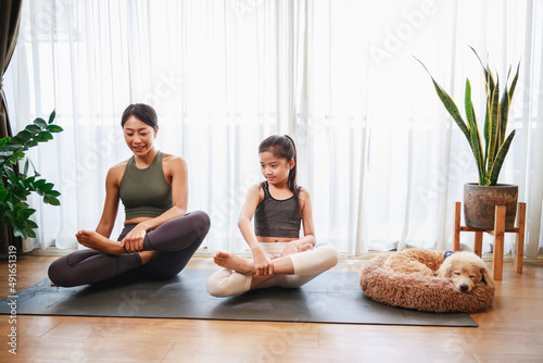 Asian young mother teaching her daughter to yoga pose and exercise together with their dog on yoga mat in living room at home.