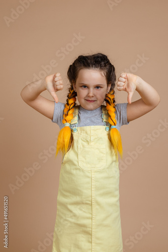 Young female with yellow kanekalon pigtails looking at camera, showing dislike signs with hands smiling wearing yellow jumpsuit and gray t-shirt on beige background.